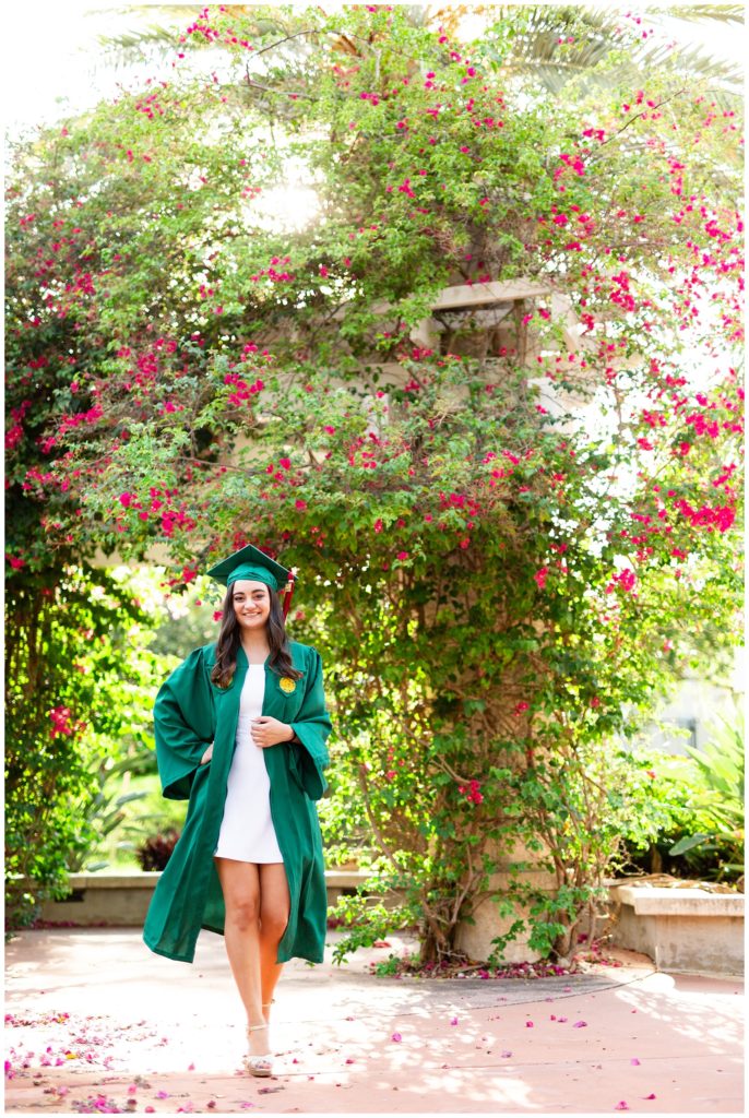 College senior walking toward camera in green cap and gown with pink flower bushes around her. 
