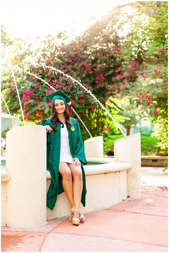 Graduate sitting by fountain in her green USF cap and gown. 