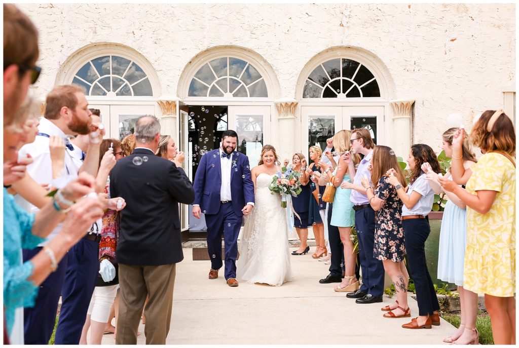 Photo of bride and groom exiting their wedding venue with guests lined up on both side outside of the wedding venue blowing bubbles as they exit