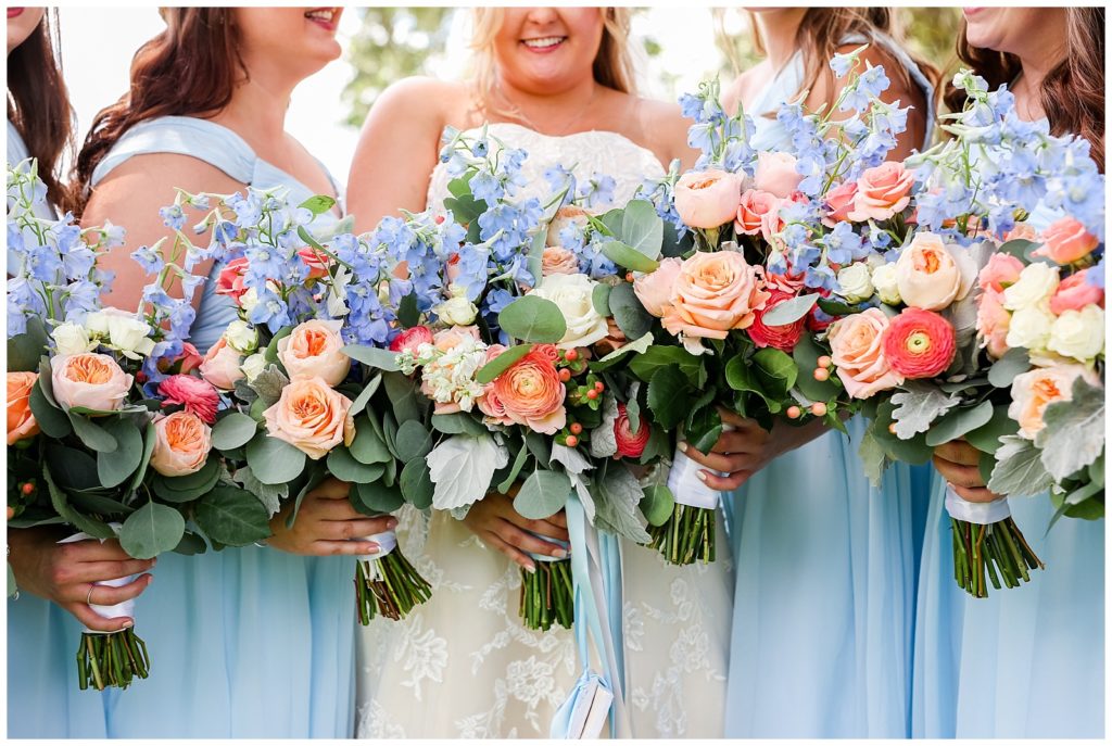 Bridesmaids holding their bouquets filled with blue, white, green and peach florals in their sky blue dresses with bride in wedding gown in the middle