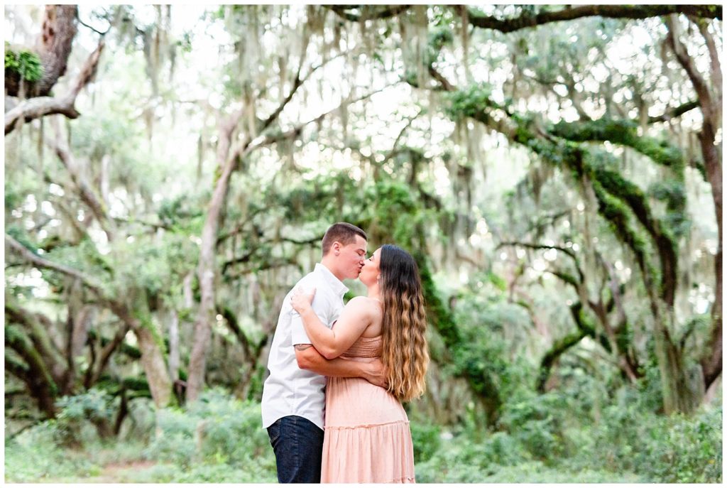 Newly engaged couple kissing under huge lush bright green trees.