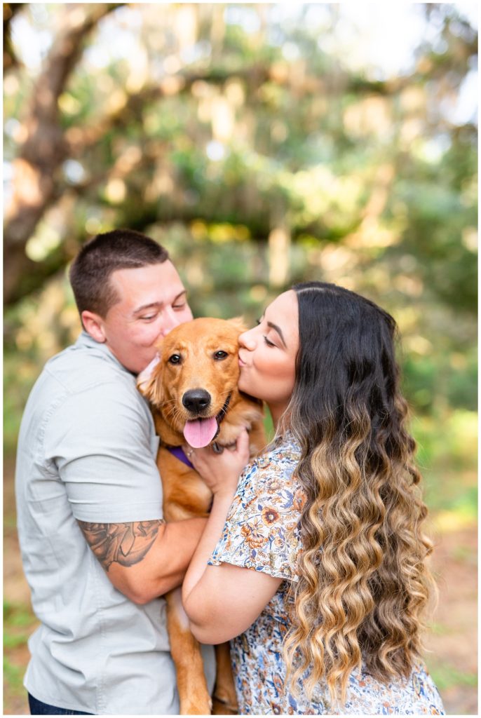 Newly engaged couple kissing their golden retriever as they hold her between them.