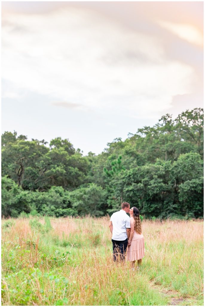 Engaged couple facing away from camera kissing in tall grass.