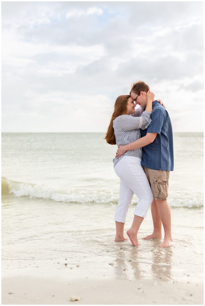 Engaged couple standing on the sand as the water touches their feet and both are looking passionately at each other in Dunedin Florida 