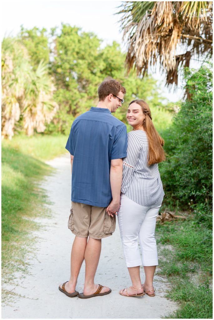 Engaged couple facing away from camera holding hands while girl looks back and smiles at the camera while both are on a white sand trail at Honeymoon Island, Dunedin Florida.