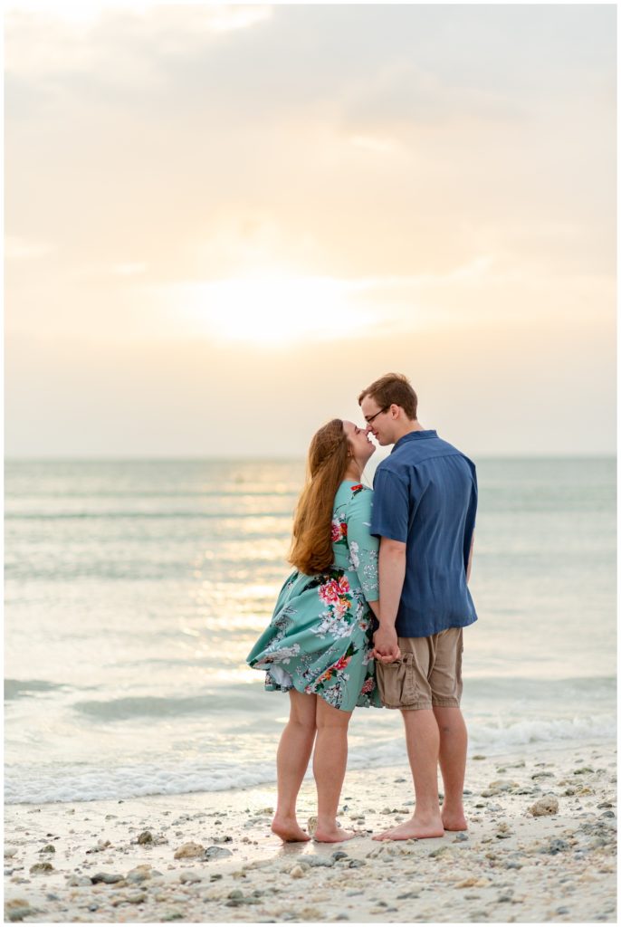 Engaged couple facing the ocean during sunset holding their hand and kissing at Honeymoon Island in Dunedin Florida. 