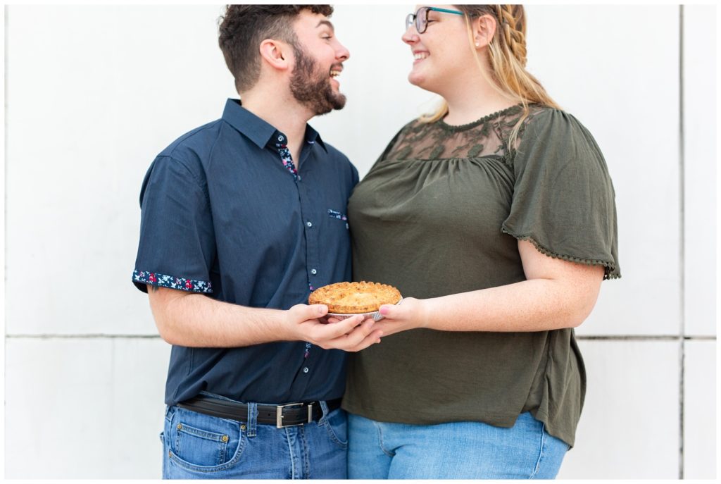 Engagement couple smiling at each other while they are holding a pie. 