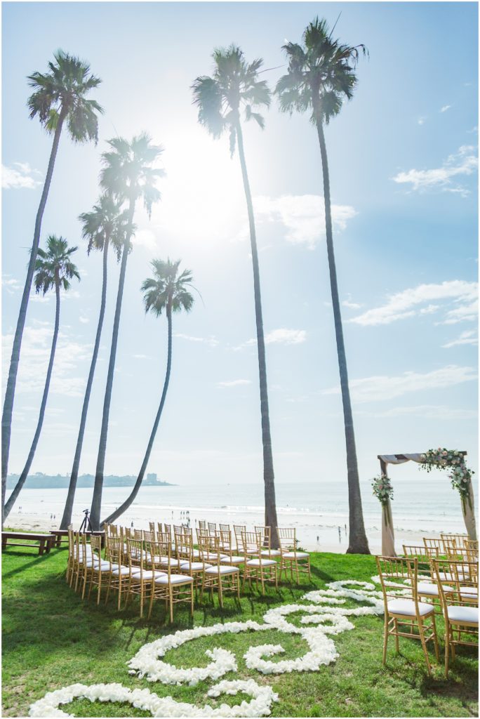 Ceremony at Scripps Seaside Forum in La Jolla California with tall palm trees, ocean in the back and rose petals swirled on lush green lawn. 