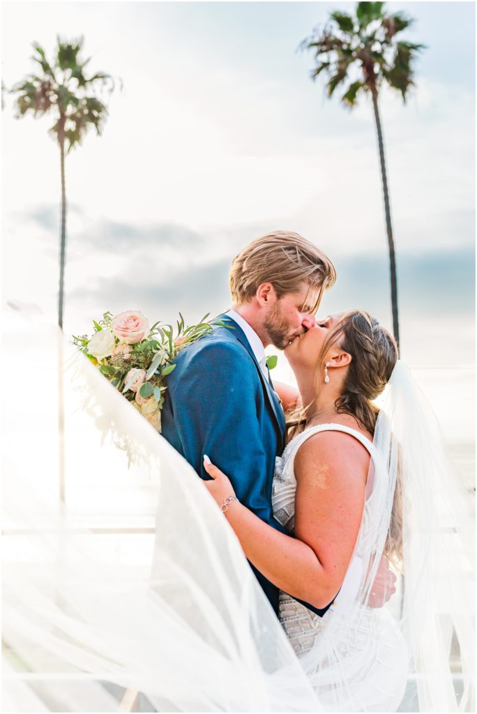 Portrait of bride and groom sharing a kiss in front of tall palm trees with veil flowing in front of the camera.