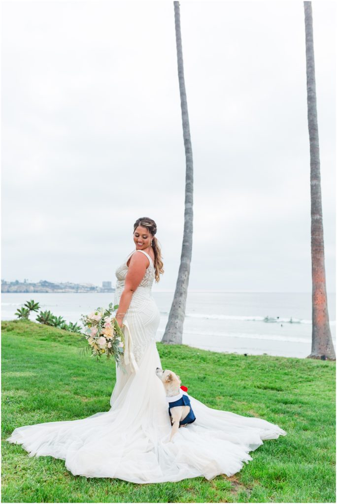 Bride's dog sitting on train of wedding dress looking up at her while she holds her bouquet and looks back at him with San Diego beach in the background.